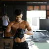 Shirtless man preparing for an intense workout session in a gym, wearing boxing gloves.