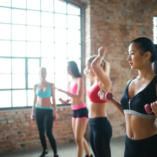 Determined pensive Asian female in black activewear holding dumbbells while training in group of other female athletes in in loft spacious gym